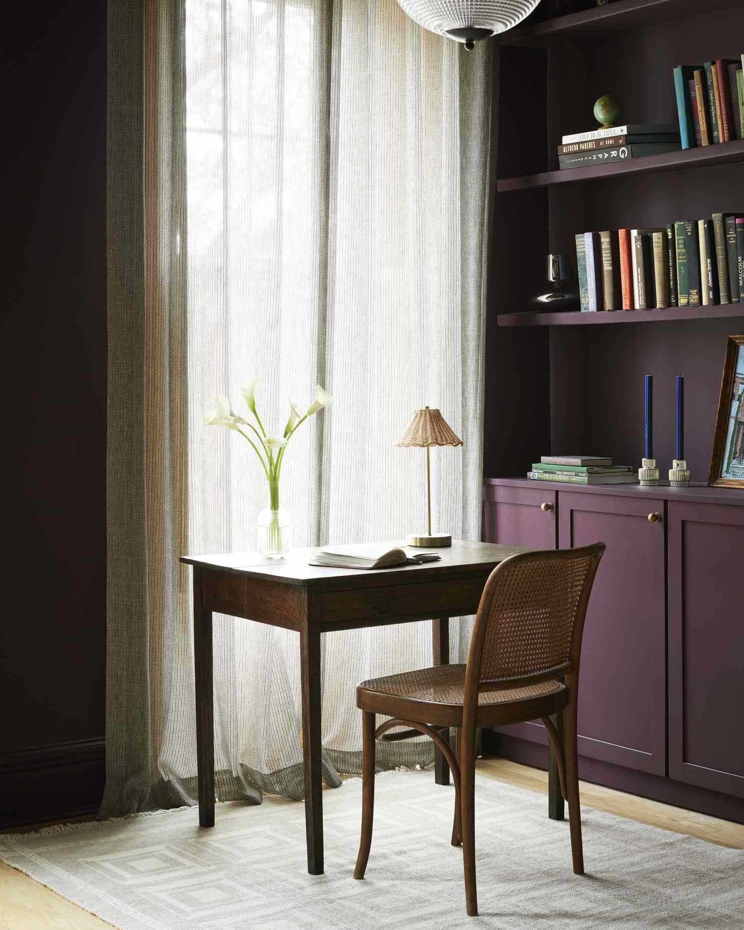 A desk and chair beside a window with sheer curtains near a bookshelf filled with books