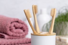 Four bamboo toothbrushes stored upright in a white holder with towels and a plant in the background