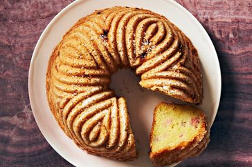 A olive-oil bundt cake on a white plate with a slice removed revealing a textured moist interior on a dark surface