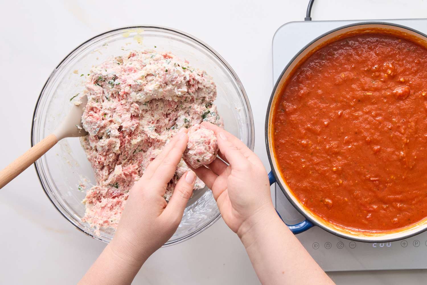 Hands shaping meatballs over a bowl of meat mixture near a pot of tomato sauce