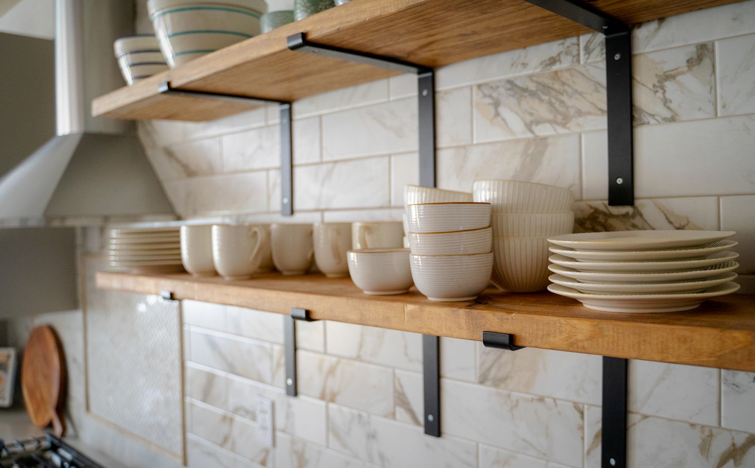 Wooden shelves in a kitchen displaying neatly arranged plates bowls and mugs with a tiled backsplash