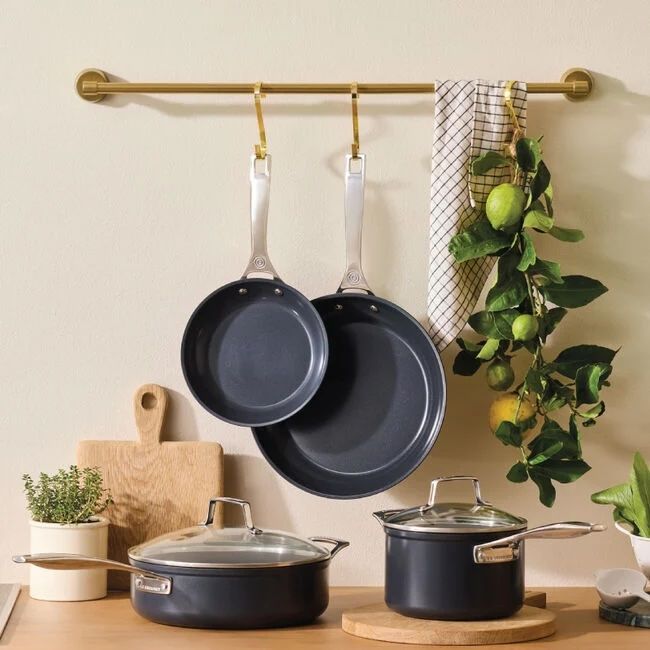 Cookware set including pans and pots with lids on a countertop with two pans hanging from a bar above