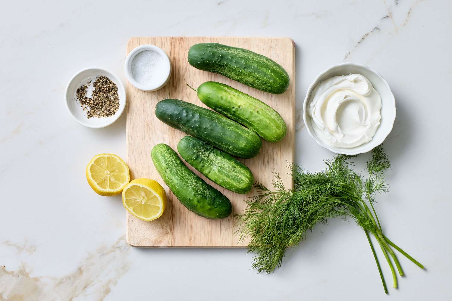 Ingredients for cucumber salad on a wooden board, including cucumbers, sour cream, dill, lemon halves, salt, and pepper