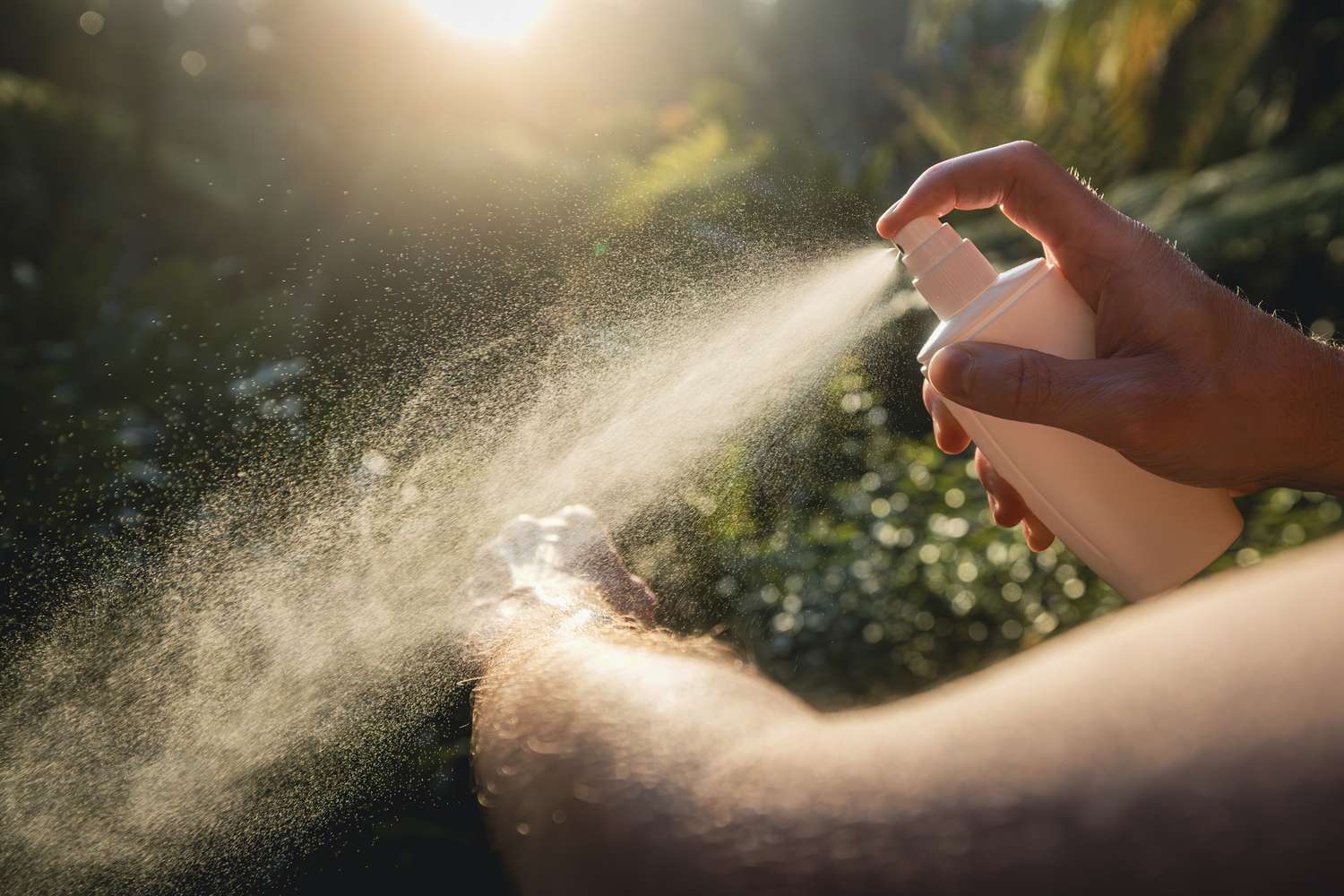 A person applying spray to their arm outdoors with sunlight in the background