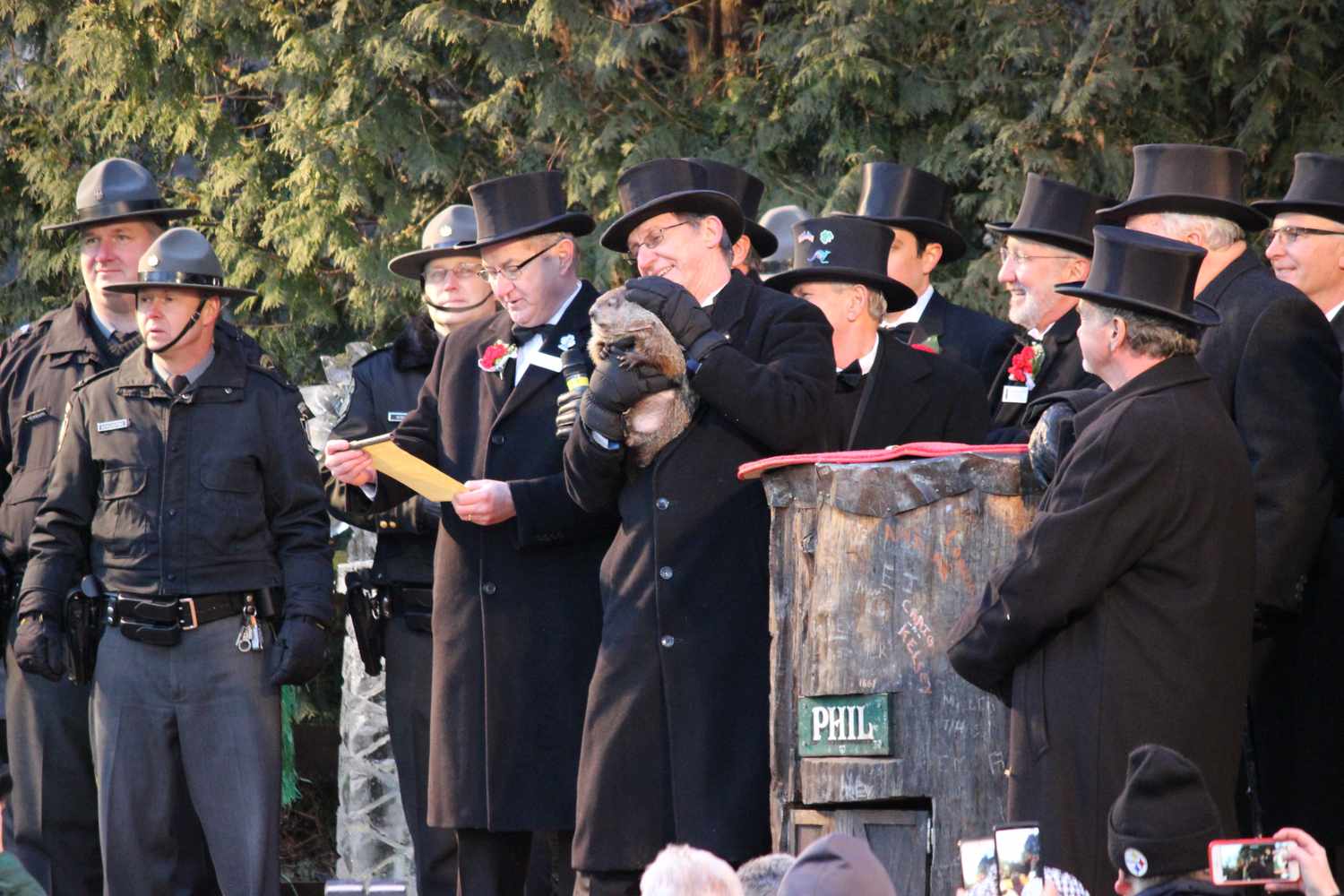 Event participants gather with a man holding a groundhog surrounding a wooden stump labeled Phil in a ceremony setting