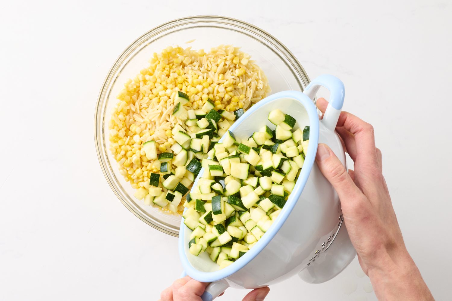 A person adding diced zucchini from a colander to a glass bowl containing cooked orzo and corn
