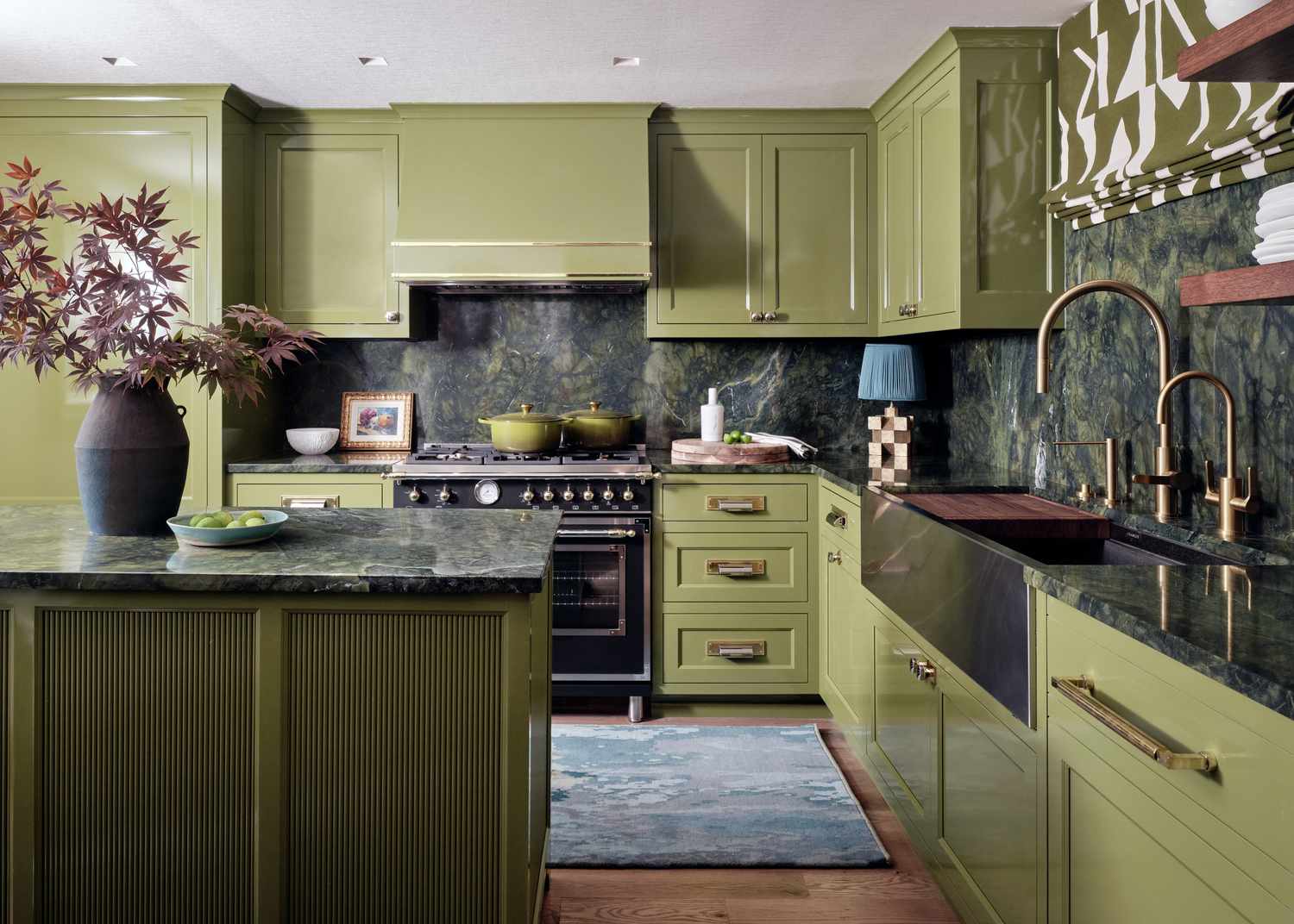 Kitchen design by Morgan Madison Design | Photography by Stephen Karlisch: glossy olive cabinetry, dramatic green marble countertops and backsplash, brass hardware, and a statement black range. A custom fluted island and patterned Roman shade add rich texture and dimension.