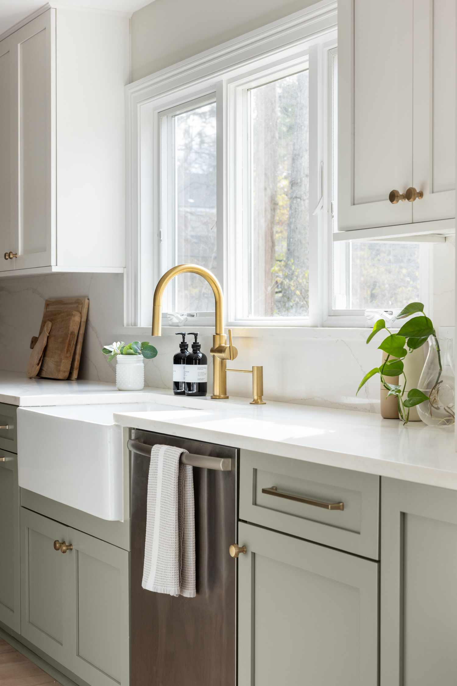 Kitchen design by Madison Hudson Designs: sage green lower cabinetry, crisp white quartz countertops, a farmhouse sink, and brass fixtures. Large windows flood the space with natural light, creating a bright and airy feel.