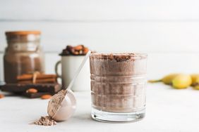 A glass of a smoothie or protein shake with a scoop of powder with additional ingredients blurred in the background