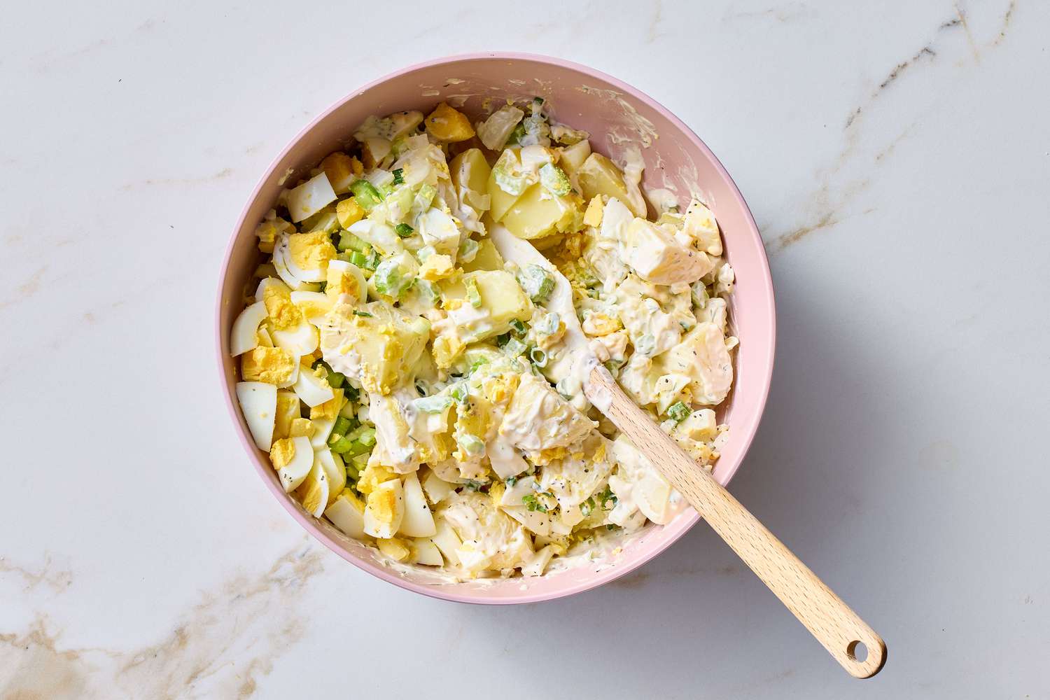 A bowl of potato salad with chopped ingredients and a mixing utensil, prepared on a light surface