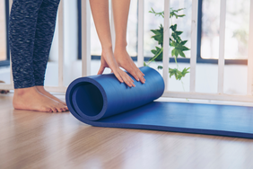 Person rolling up a yoga mat on a wooden floor indoors with a railing and plant in the background