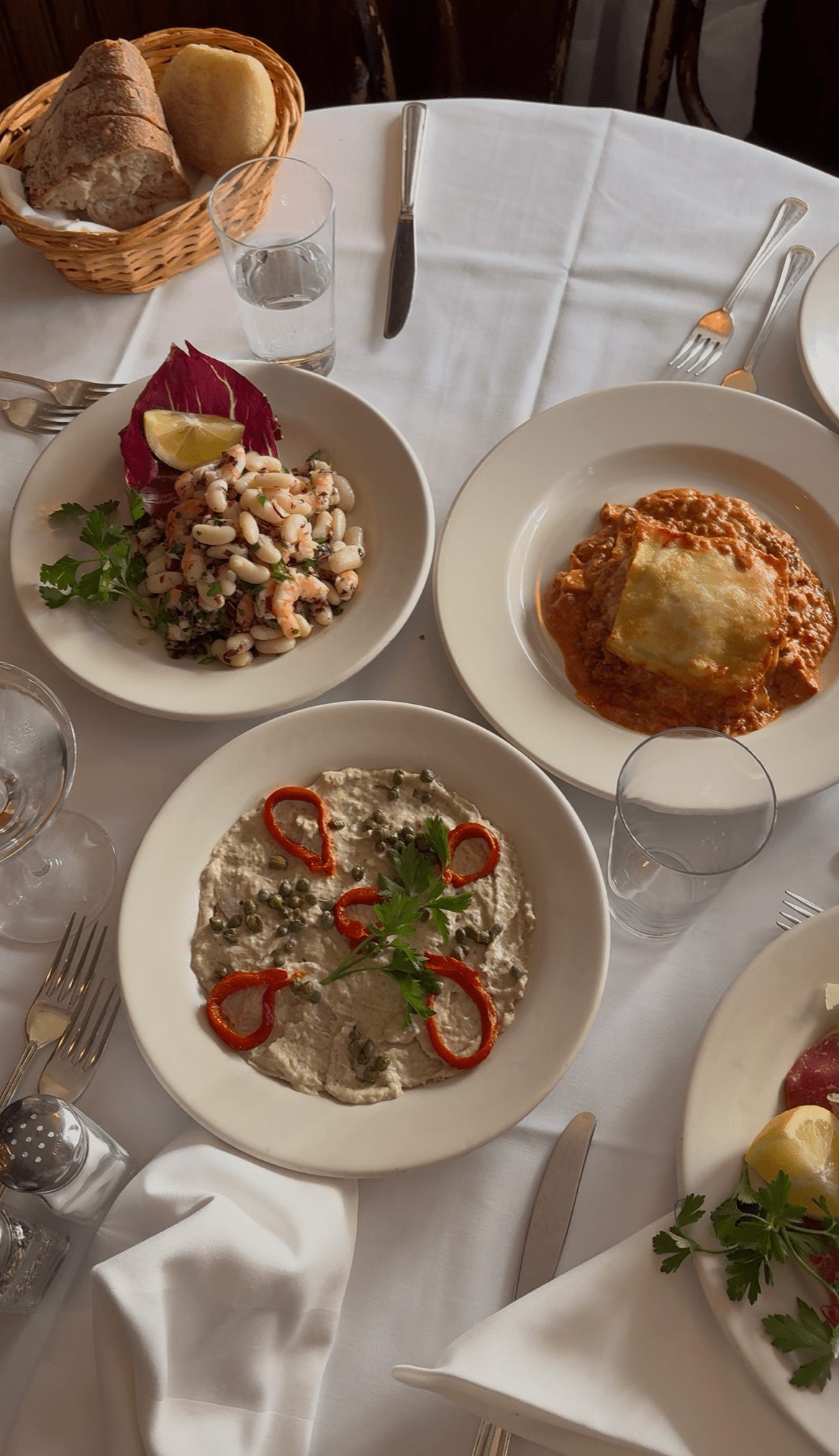 Plated dinner featuring bean salad, lasagna, bread basket, and veal. 