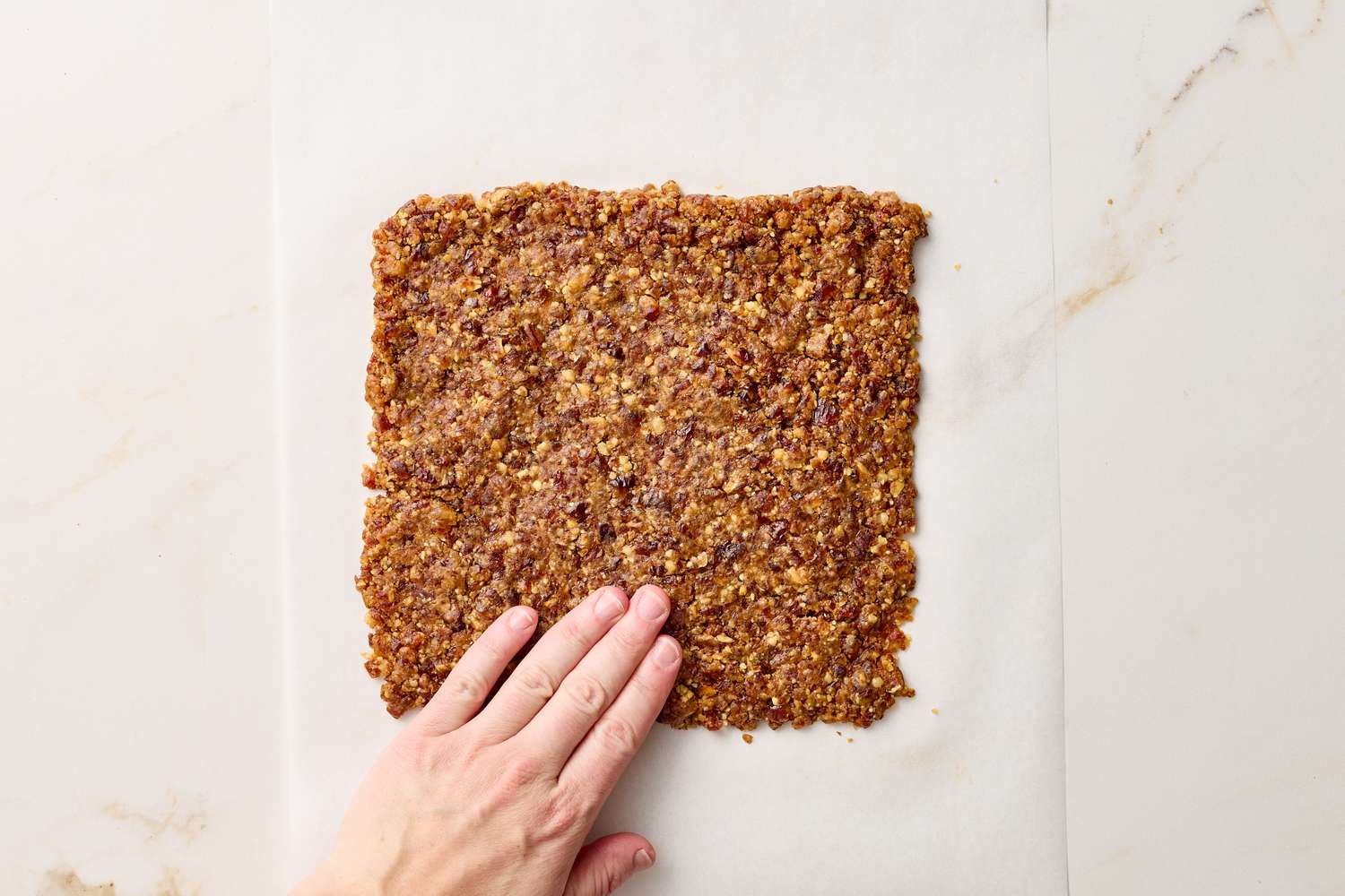 A hand pressing down a rectangular slab of dark chocolate walnut date bar mixture on parchment paper