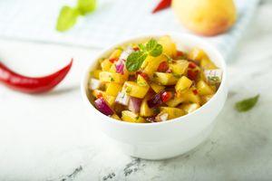 A bowl containing diced peach and vegetable salsa garnished with a mint leaf with a chili pepper and peach in the background