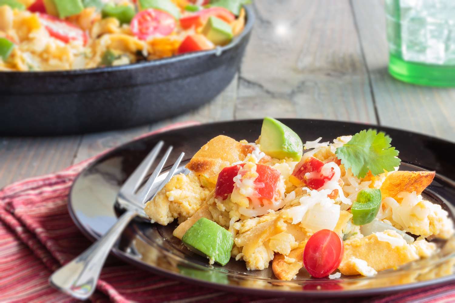 A plate of food with a fork consisting of scrambled eggs cherry tomatoes avocado cilantro and other toppings served on a dark plate with additional food in the background