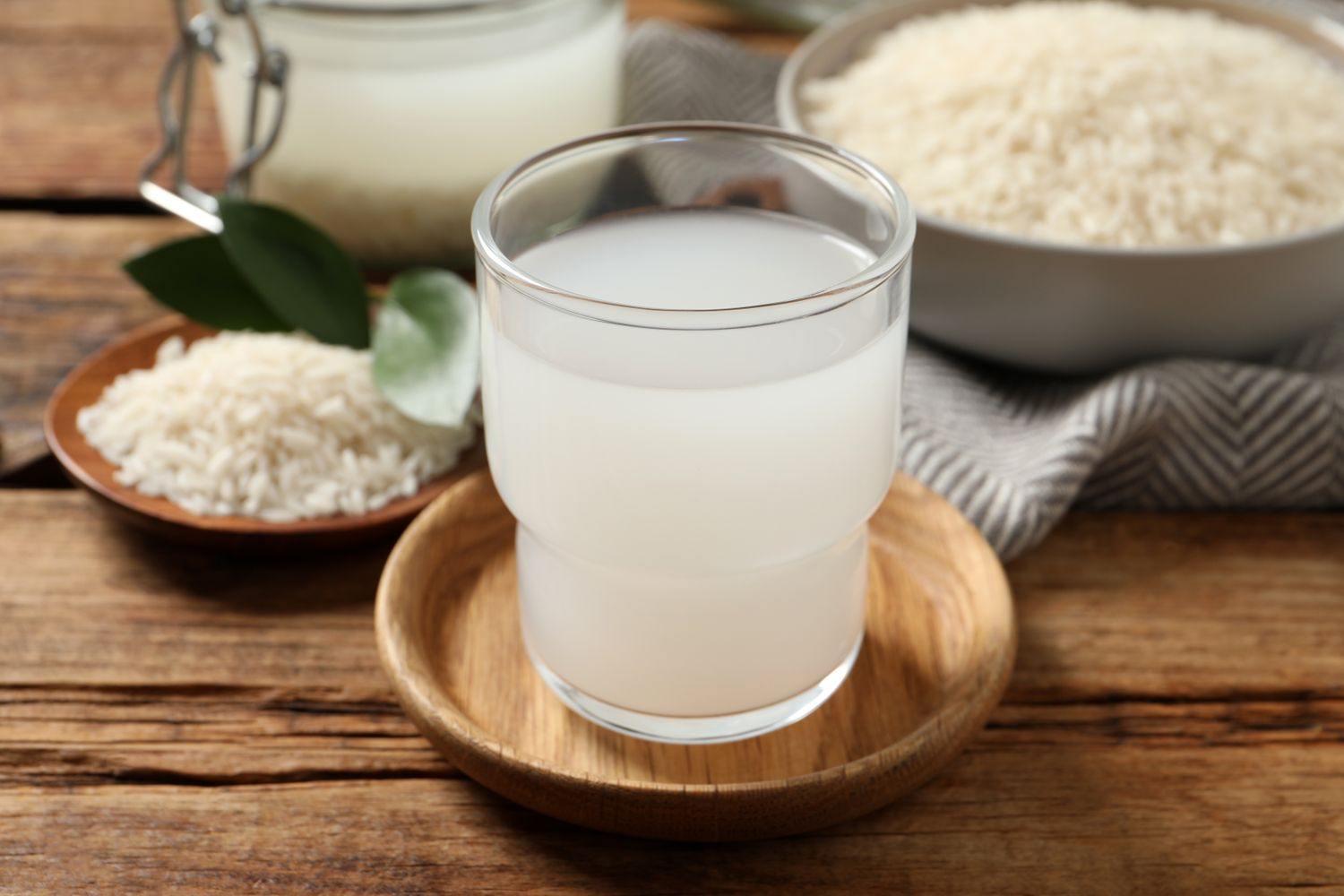 A glass of rice water on a wooden tray beside raw rice and a bowl of cooked rice