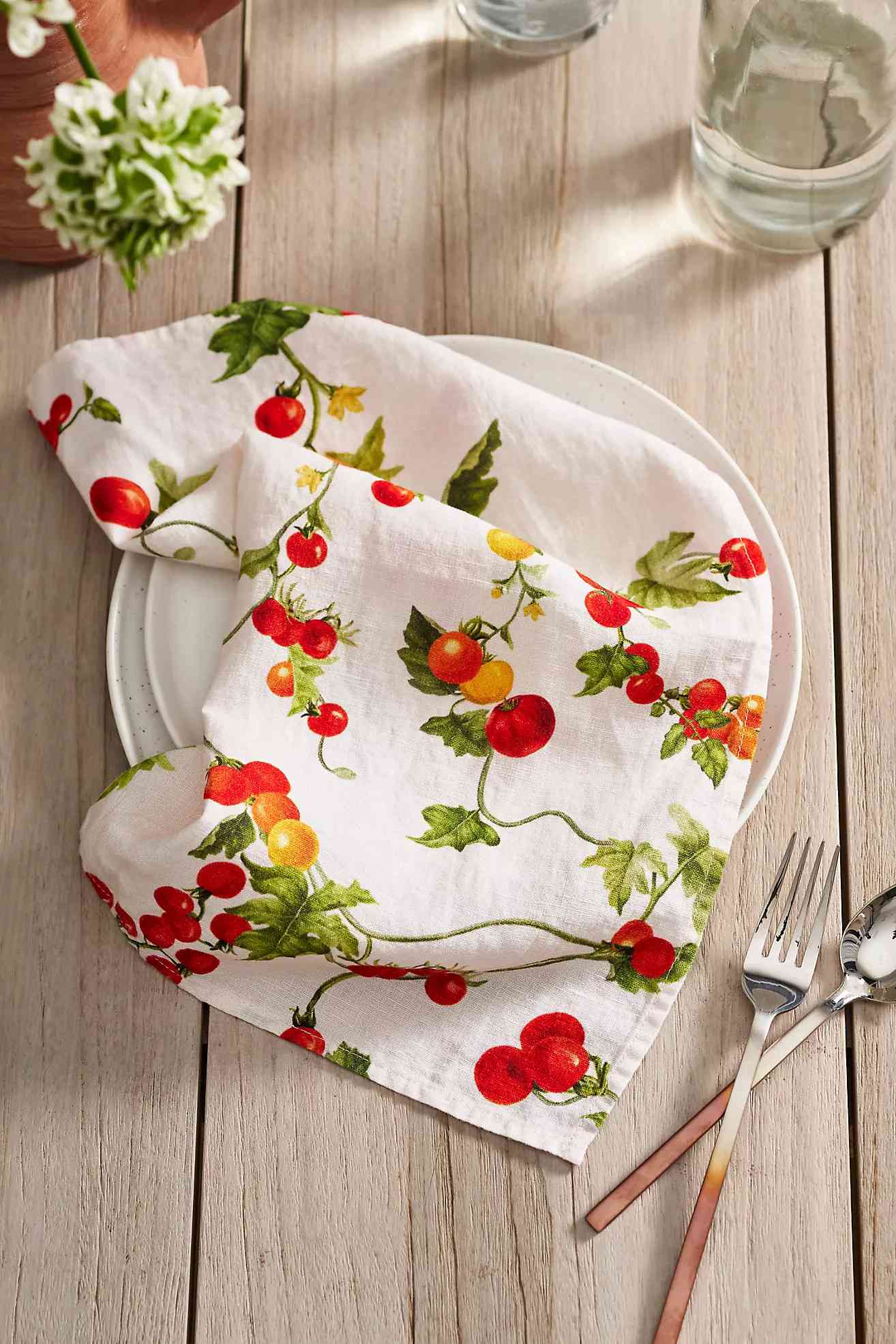A white plate on a wooden table with a tomato-patterned cloth draped over it, along with utensils and a glass