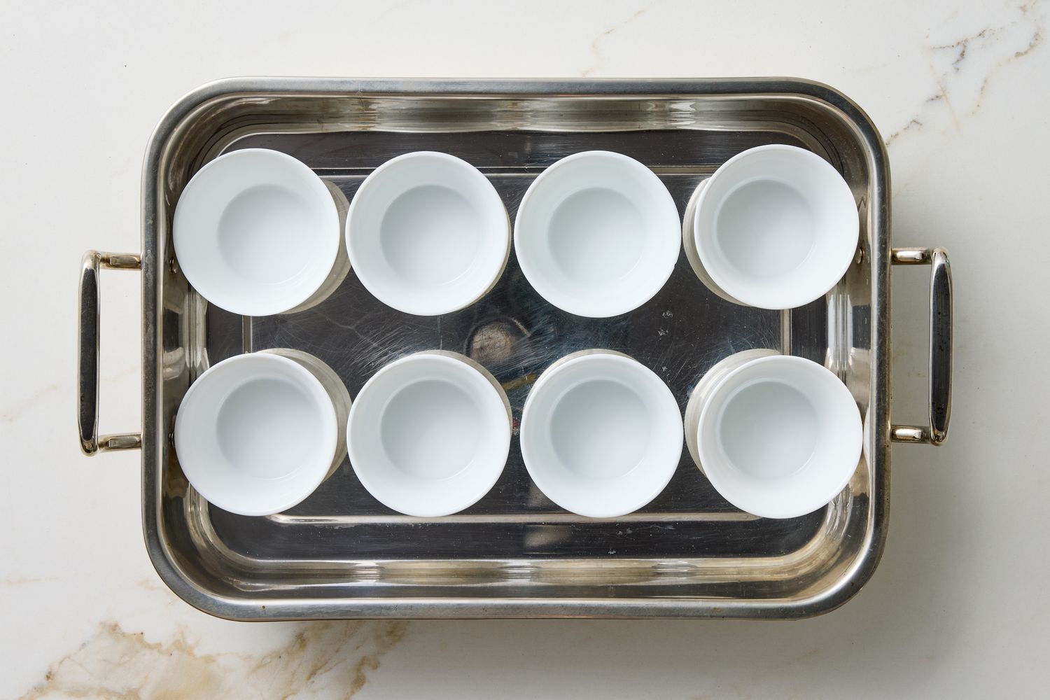 A baking tray holding eight white ramekins on a marble surface
