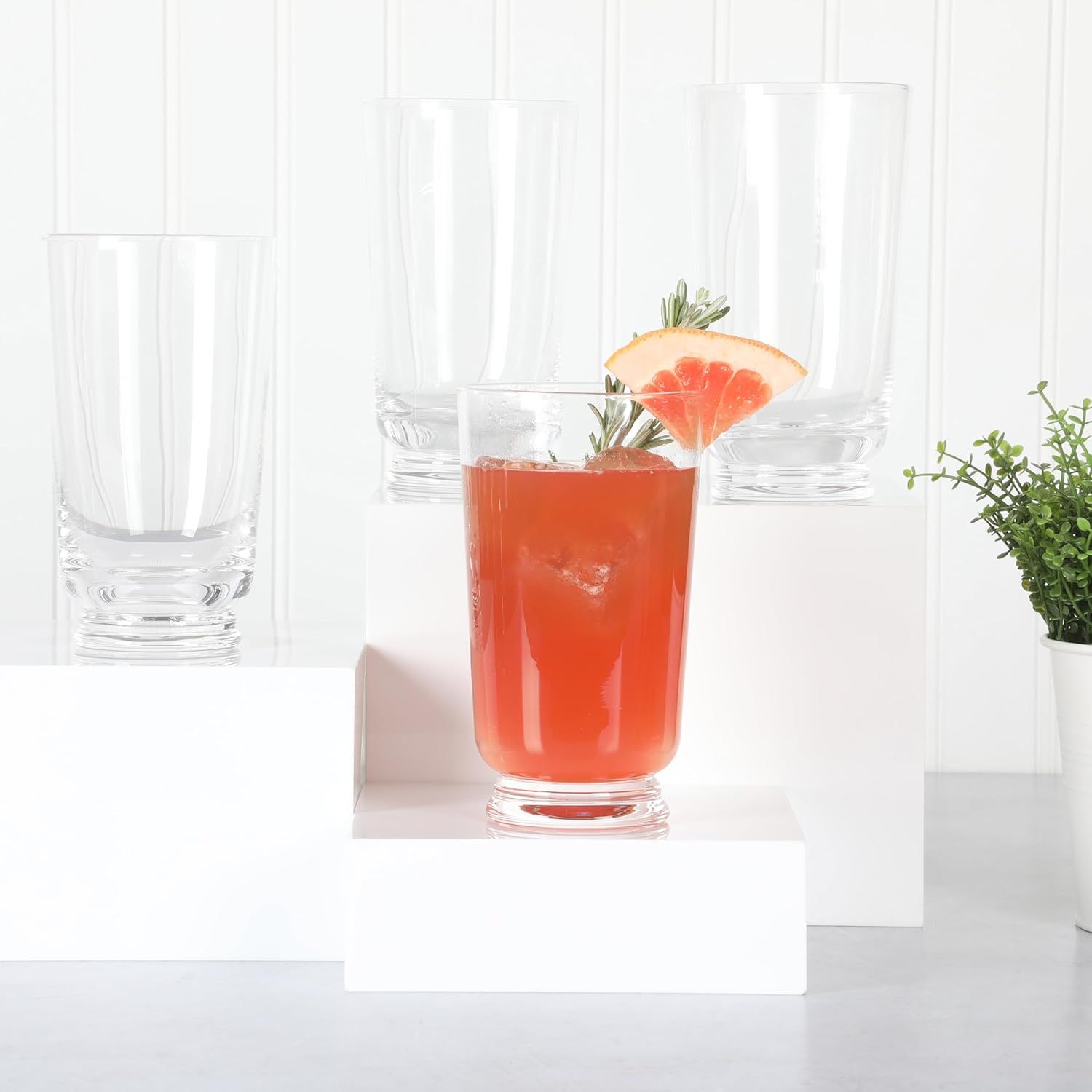A glass of redorange drink with garnish displayed alongside empty glasses on a white background
