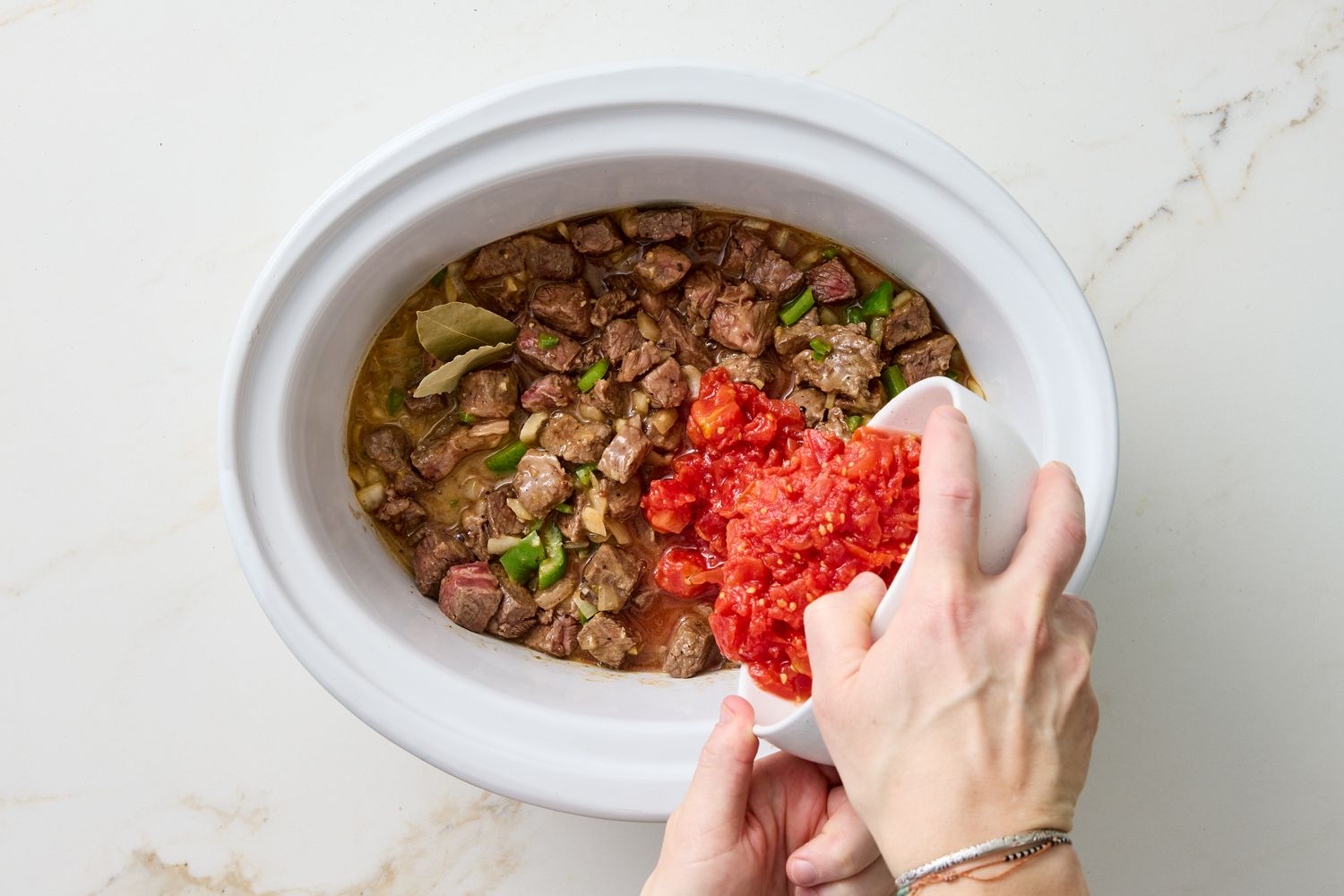 Closeup of hands adding diced tomatoes to a slow cooker with simmering beef and vegetables