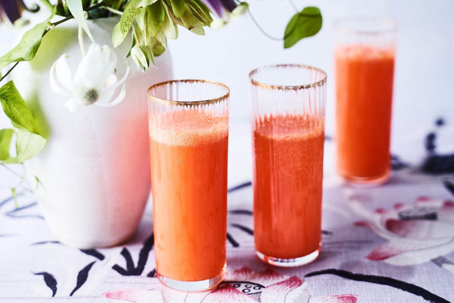 Three glasses of an orange beverage on a floralpatterned surface next to a white vase with flowers and greenery