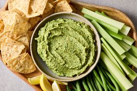 A bowl of guacamole hummus surrounded by tortilla chips celery sticks green beans and lemon wedges