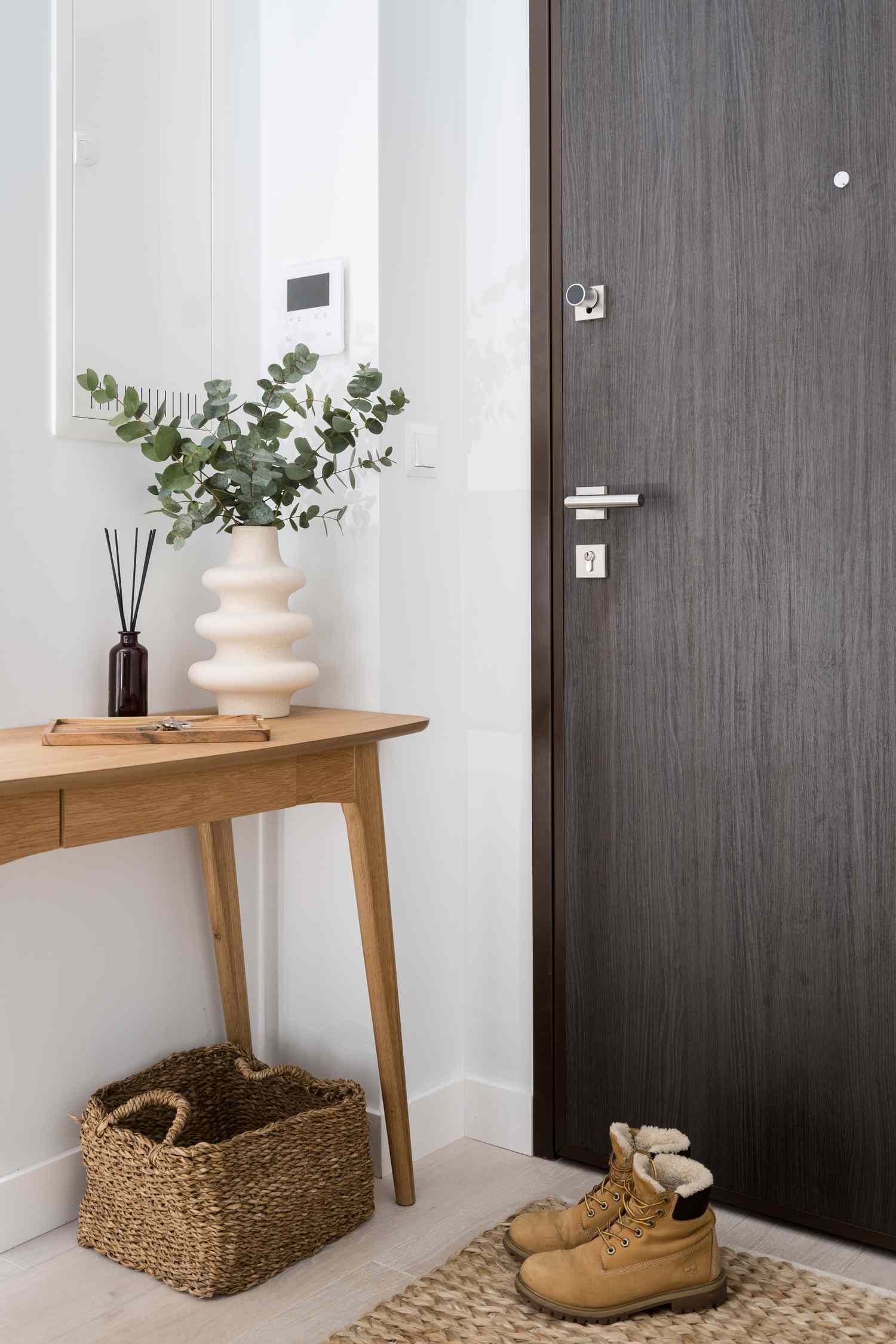 Entryway with a dark wood door a wooden side table with a decorative vase and greenery wicker basket below and a pair of boots on a rug
