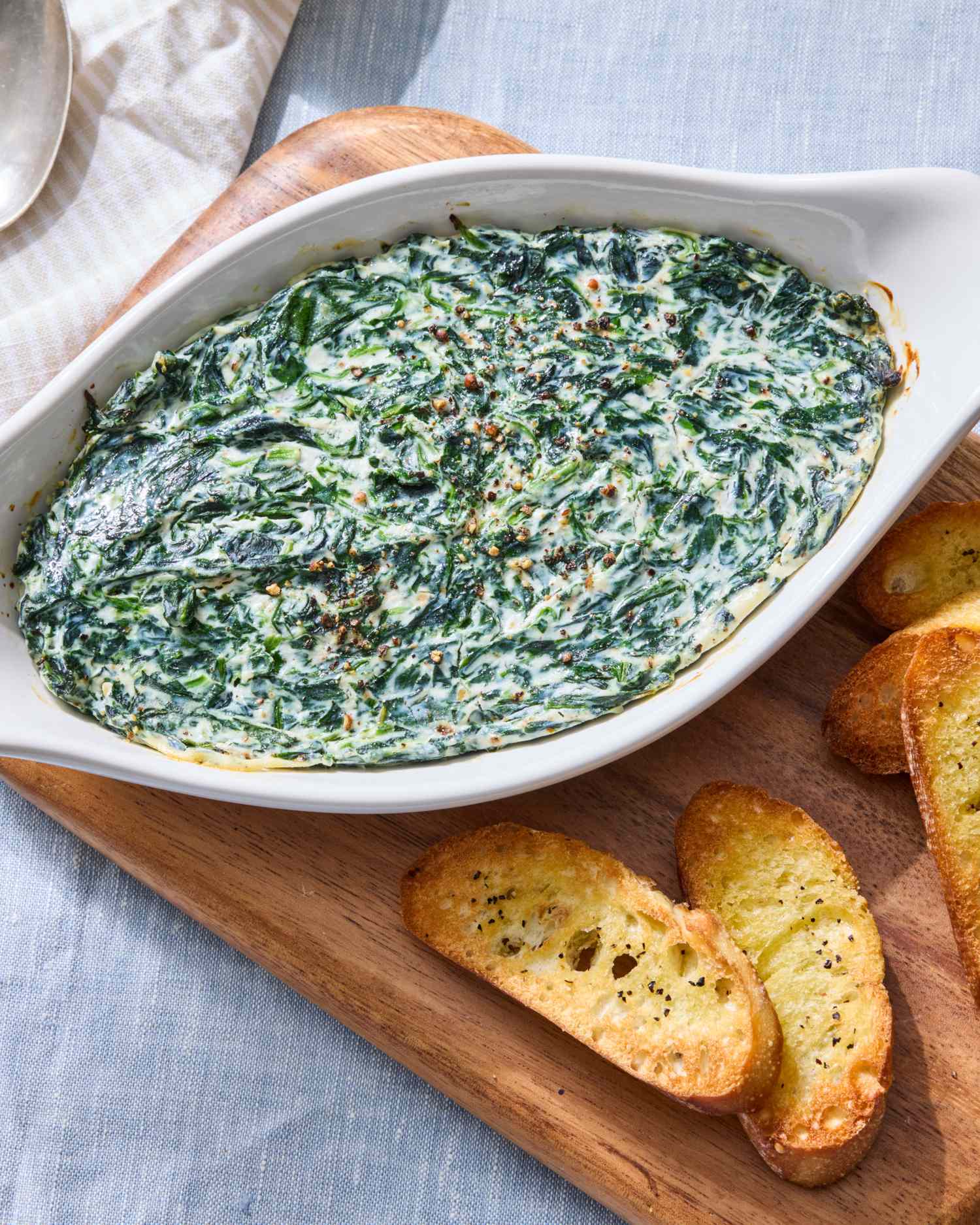 A serving dish of creamy spinach dip on a wooden board accompanied by toasted bread slices
