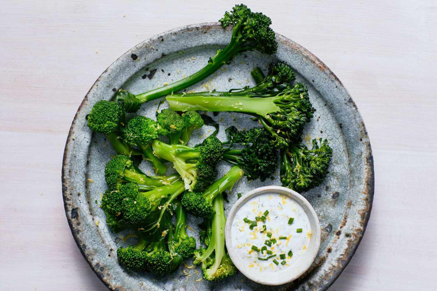 A plate with steamed broccoli florets and a small dish of ranch dip topped with chives