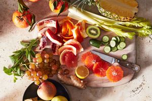 A variety of cut fruits and vegetables on a wooden board, including apples, citrus, celery, pineapple, carrots, grapes, and cucumber