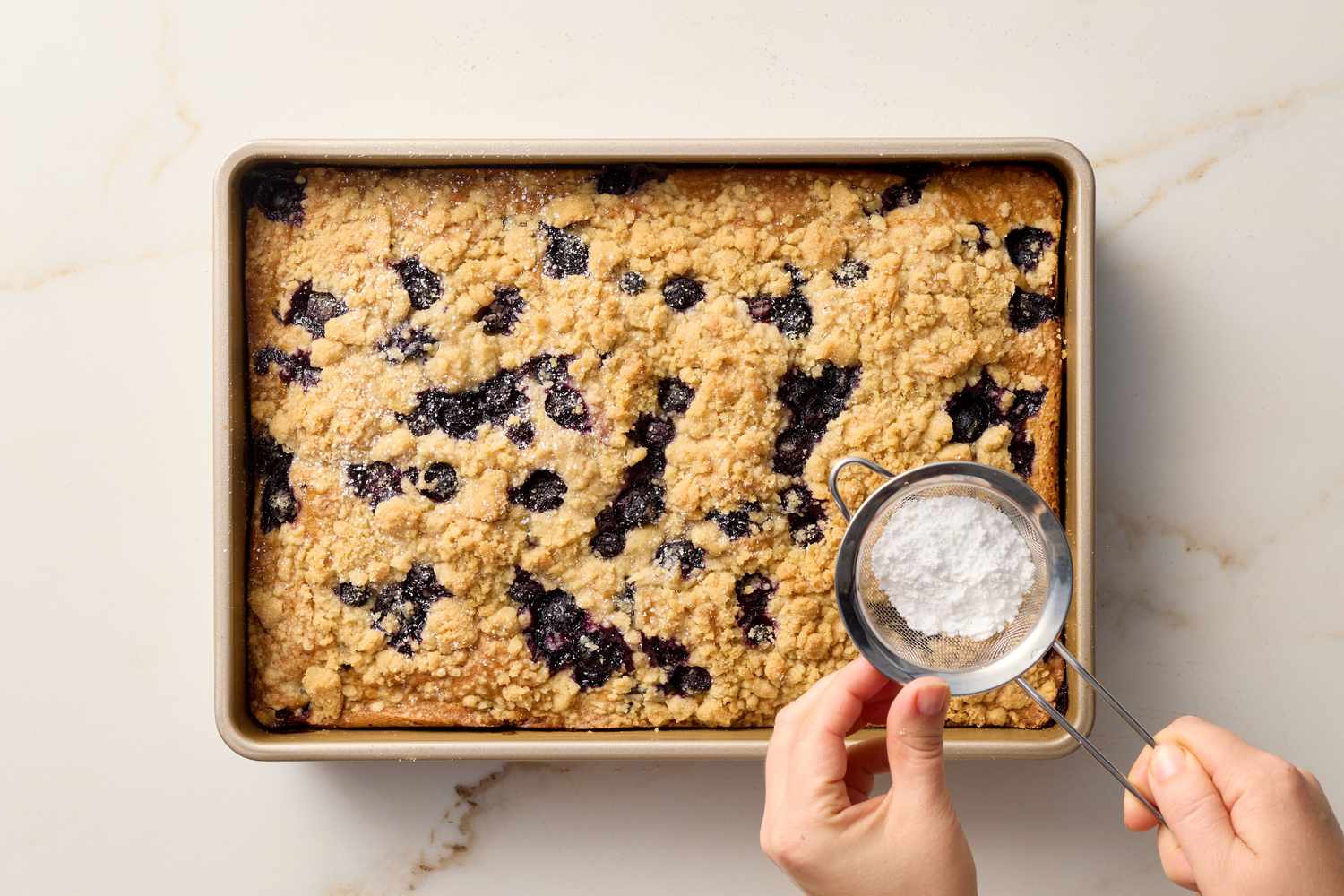 A tray of blueberry crumb cake is topped with powdered sugar using a small sieve by a person's hand