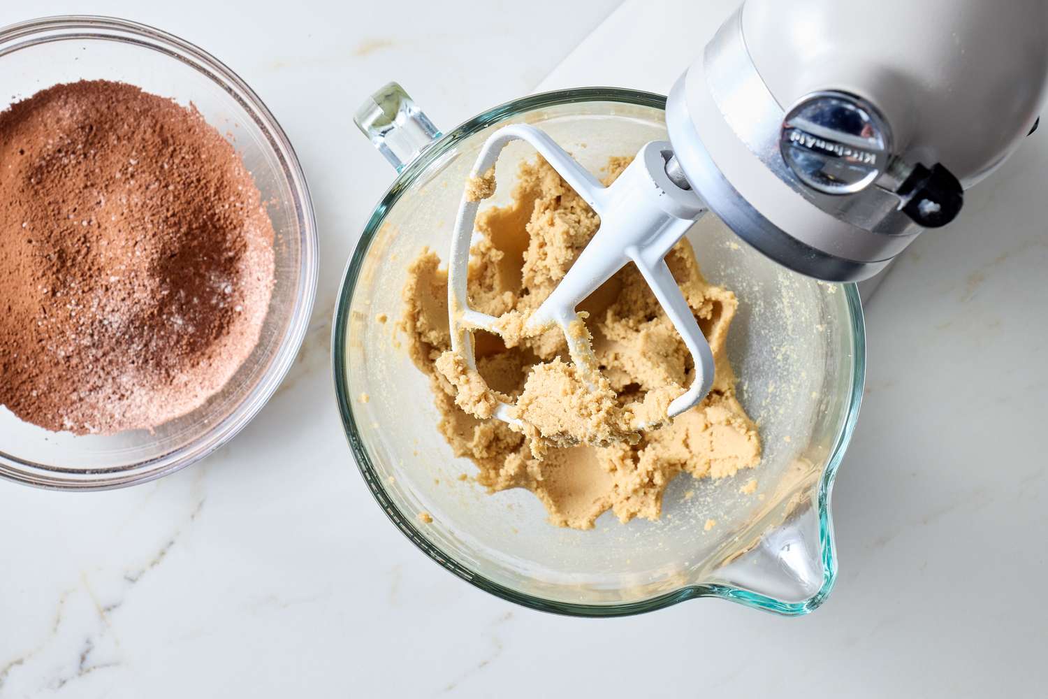 A mixing bowl with dough and a stand mixer attachment next to a bowl of cocoa powder and sugar on a counter