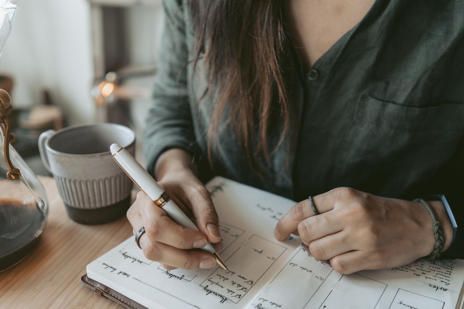 A person writing in a notebook with a pen a mug and desk items in view