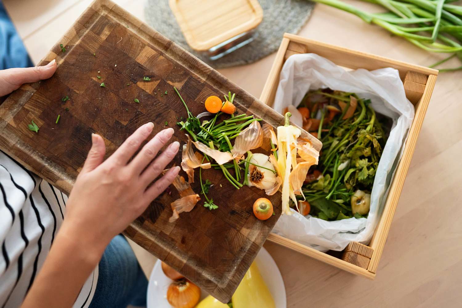 Person discarding vegetable scraps from a cutting board into a compost bin overhead view of kitchen setup