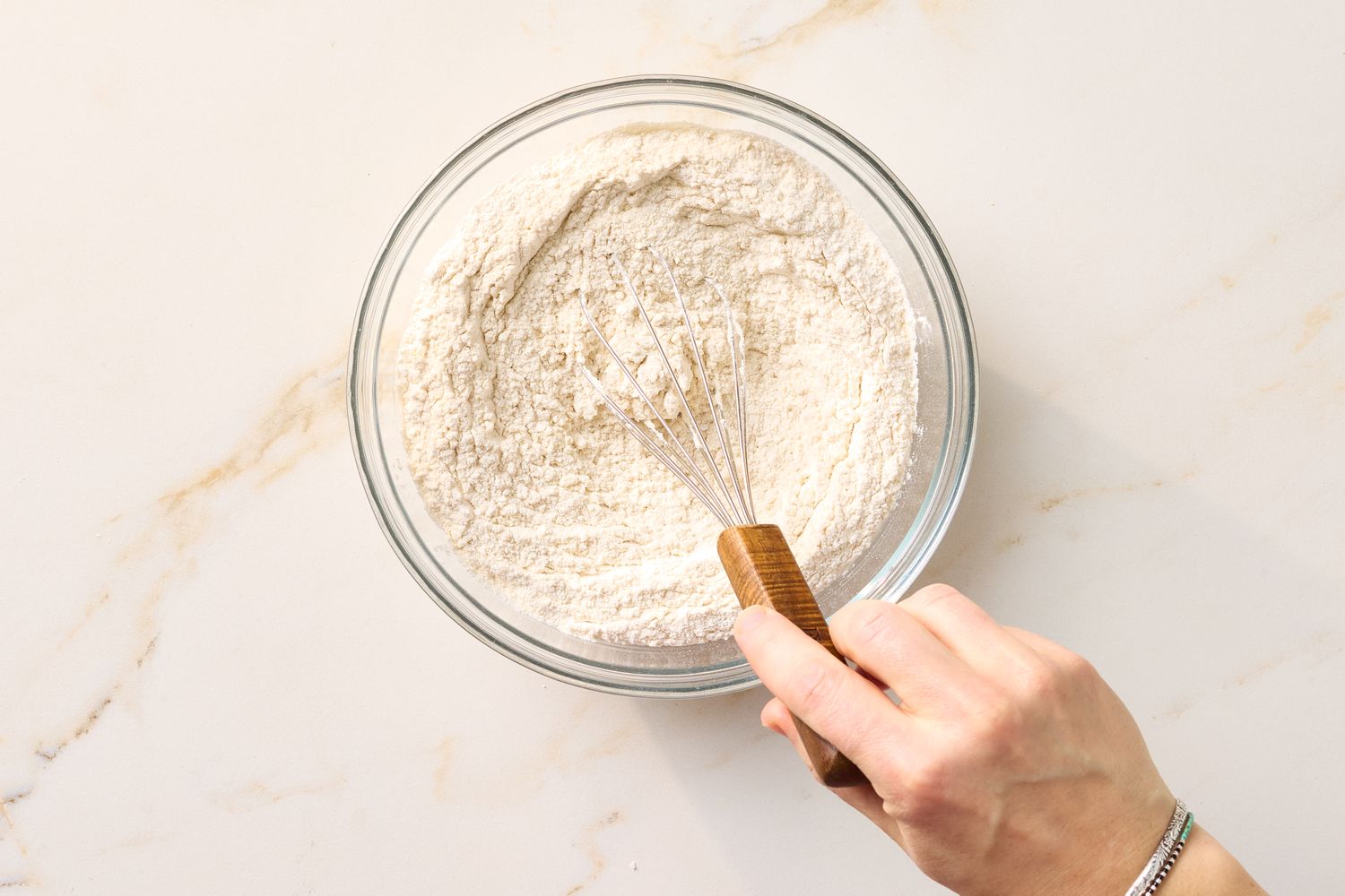 Person whisking flour in a glass bowl on a countertop preparing baking ingredients