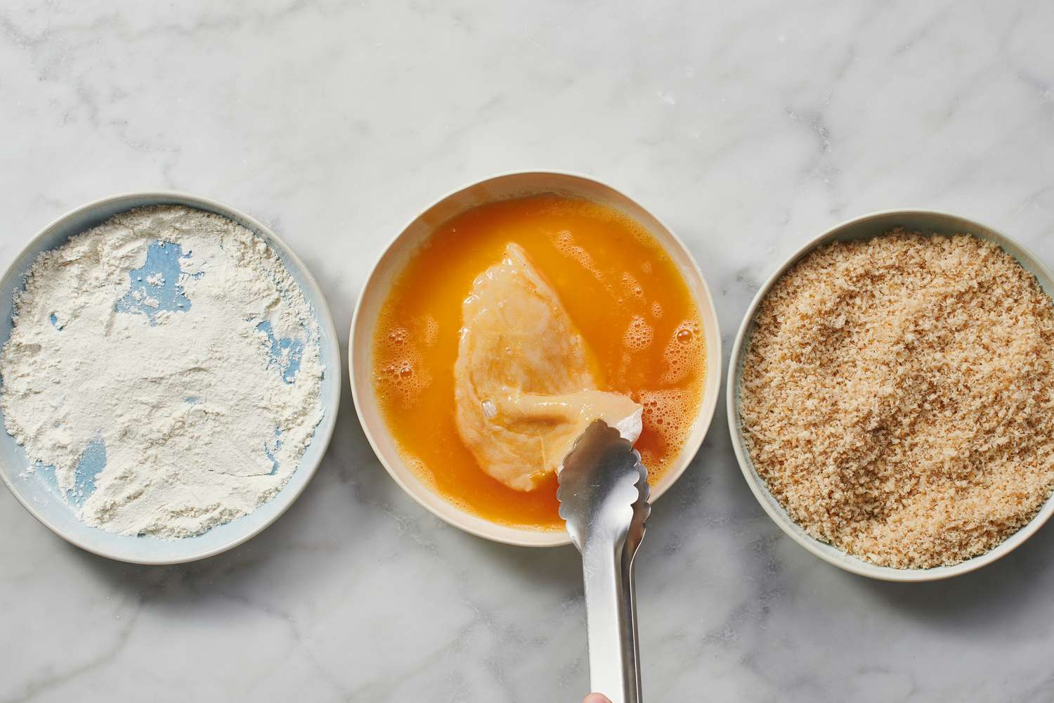 tongs holding chicken breast being coated in beaten egg in dish