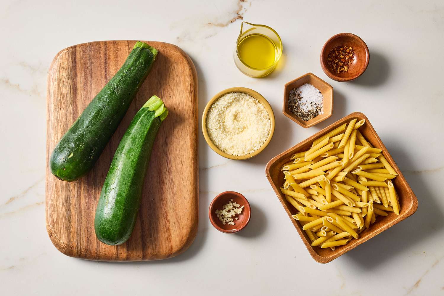 Ingredients including zucchini penne pasta grated cheese olive oil and seasonings arranged on a surface