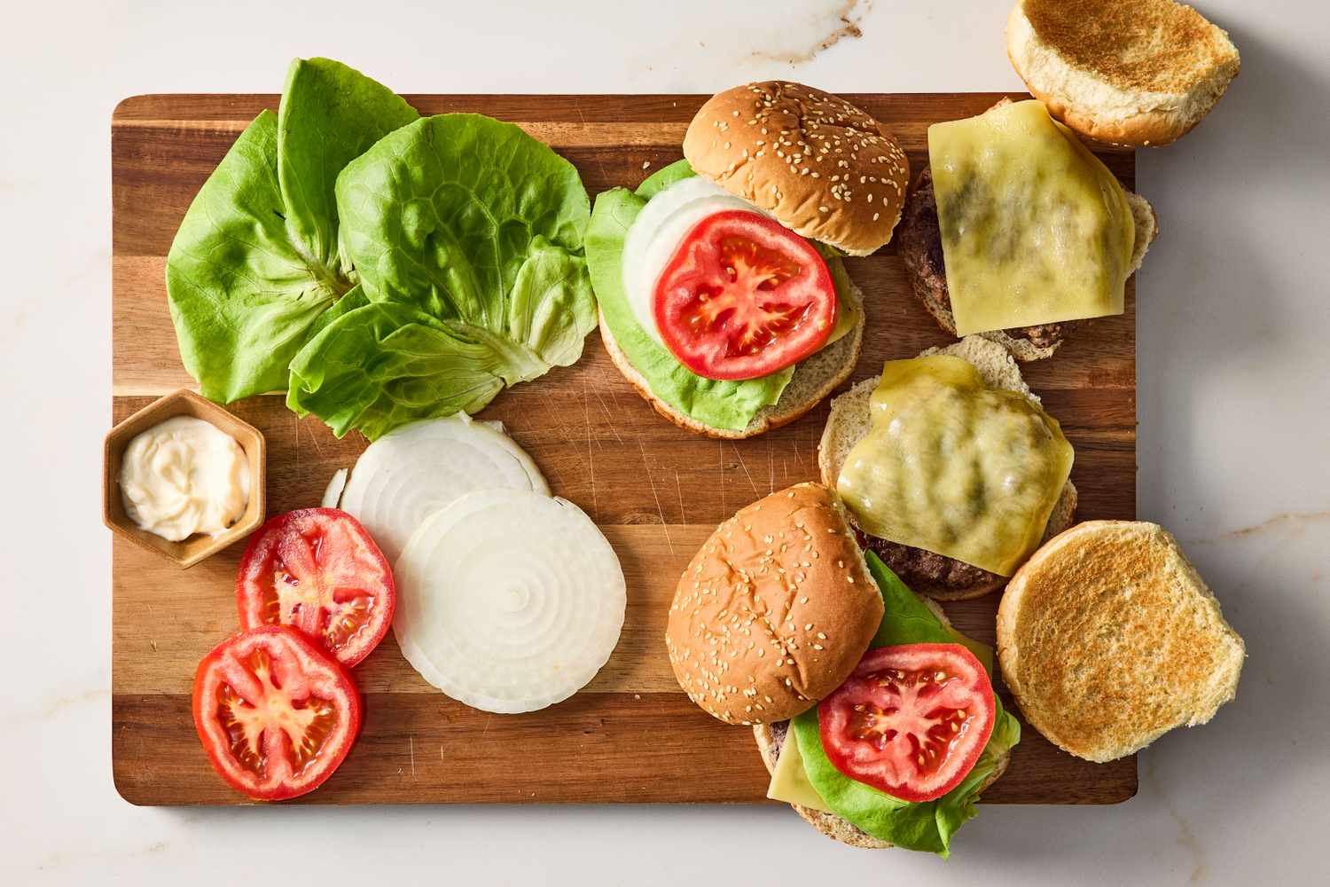 Ingredients for burgers on a cutting board including buns lettuce tomato slices onion slices and cheesecovered patties