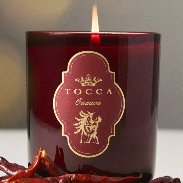 A lit Tocca Oaxaca candle with dried chili peppers on a surface in front of it