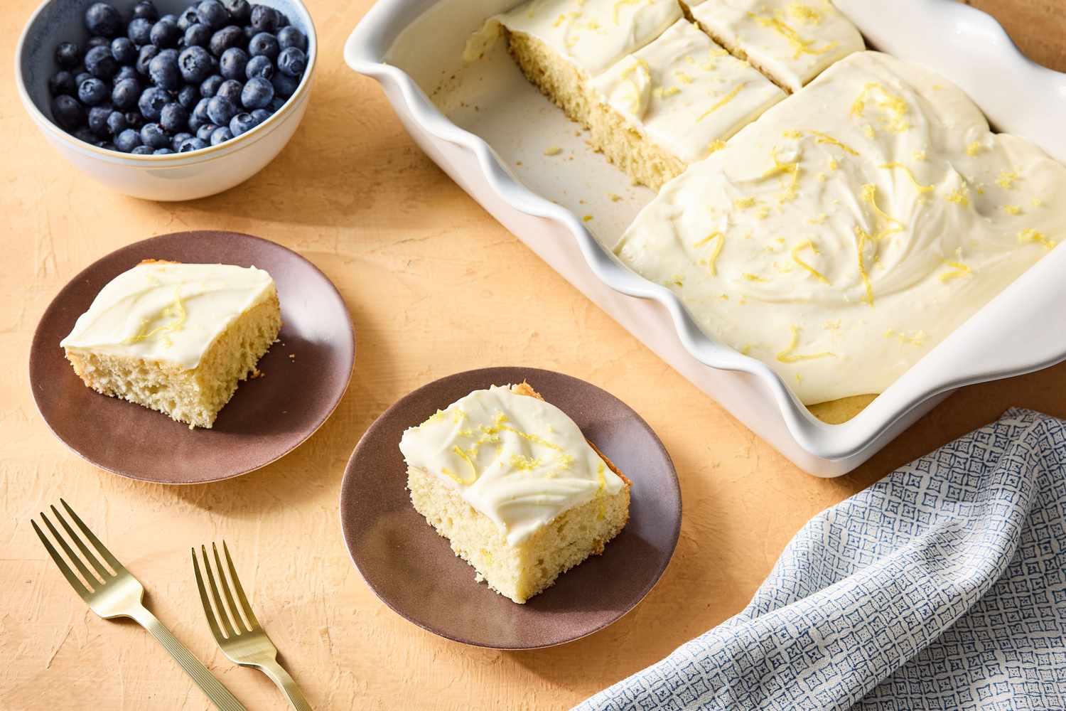 Lemon cream cheese frosted sheet cake with two servings on plates blueberries in a bowl nearby