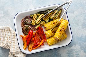 grilled corn, zucchini, onions, and bell peppers in white enamel tray with tongs.