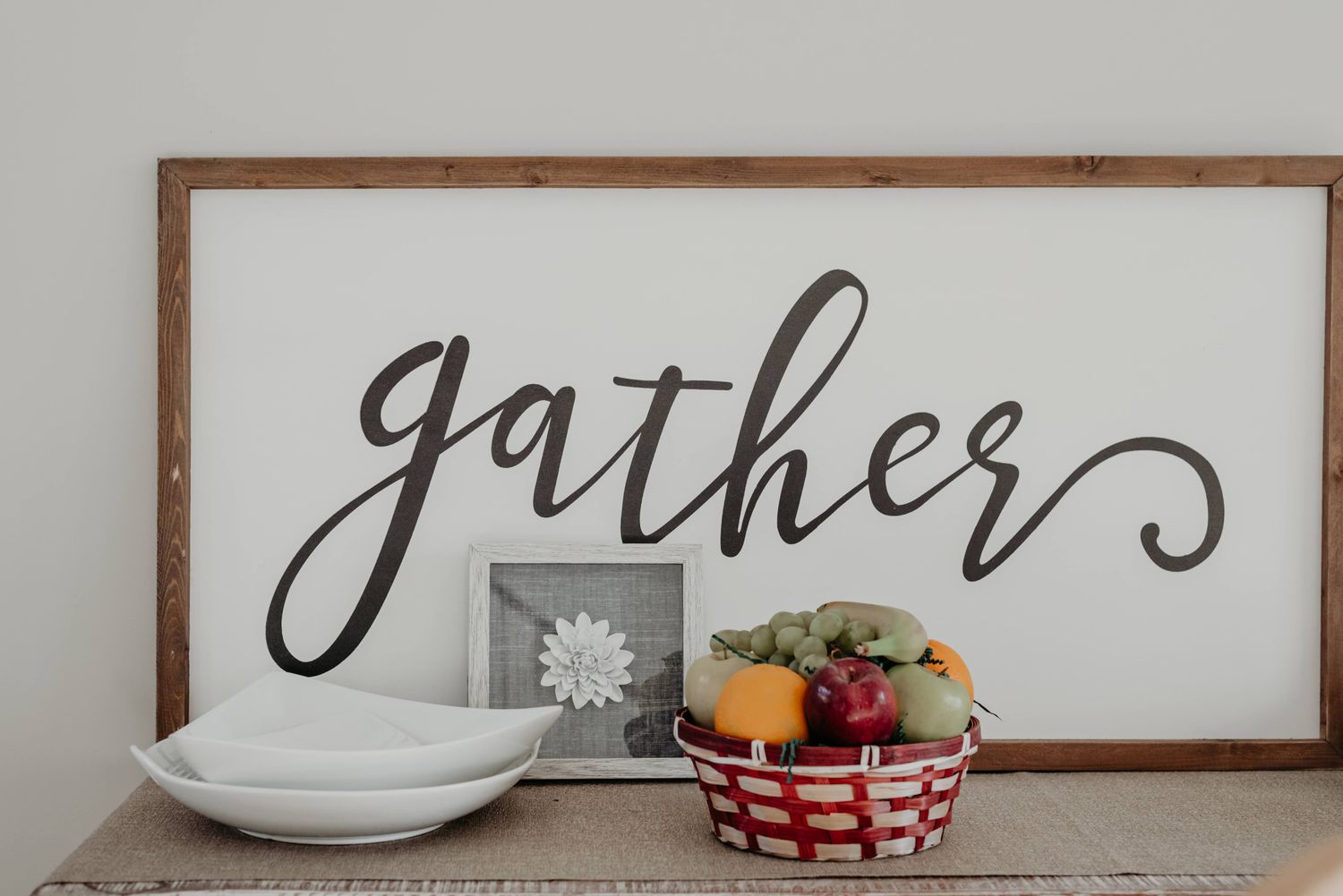 A decorative sign with the word gather displayed above a basket of fruit and some plates on a table