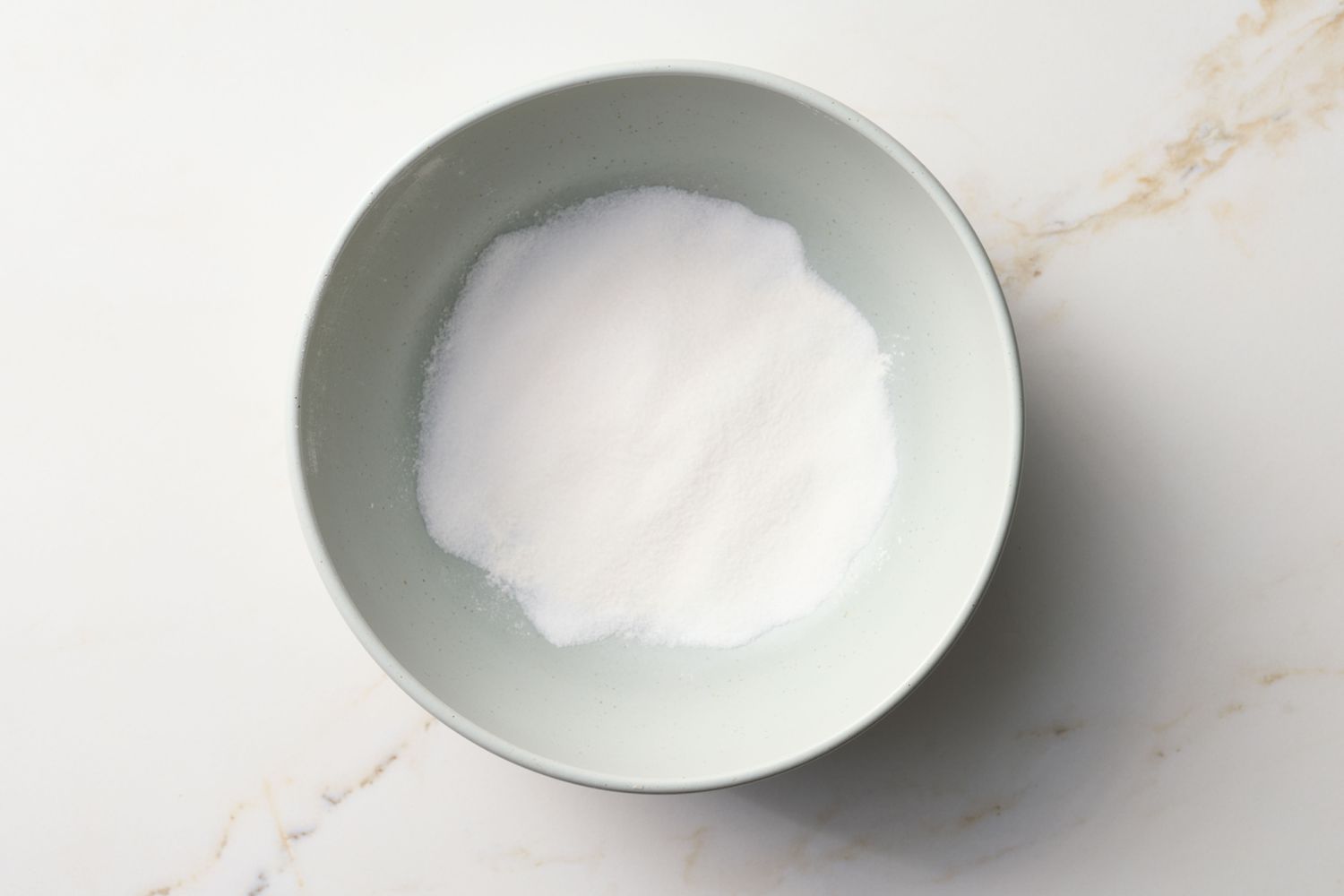 A bowl containing a white powdered substance, possibly sugar or flour, on a light marble surface