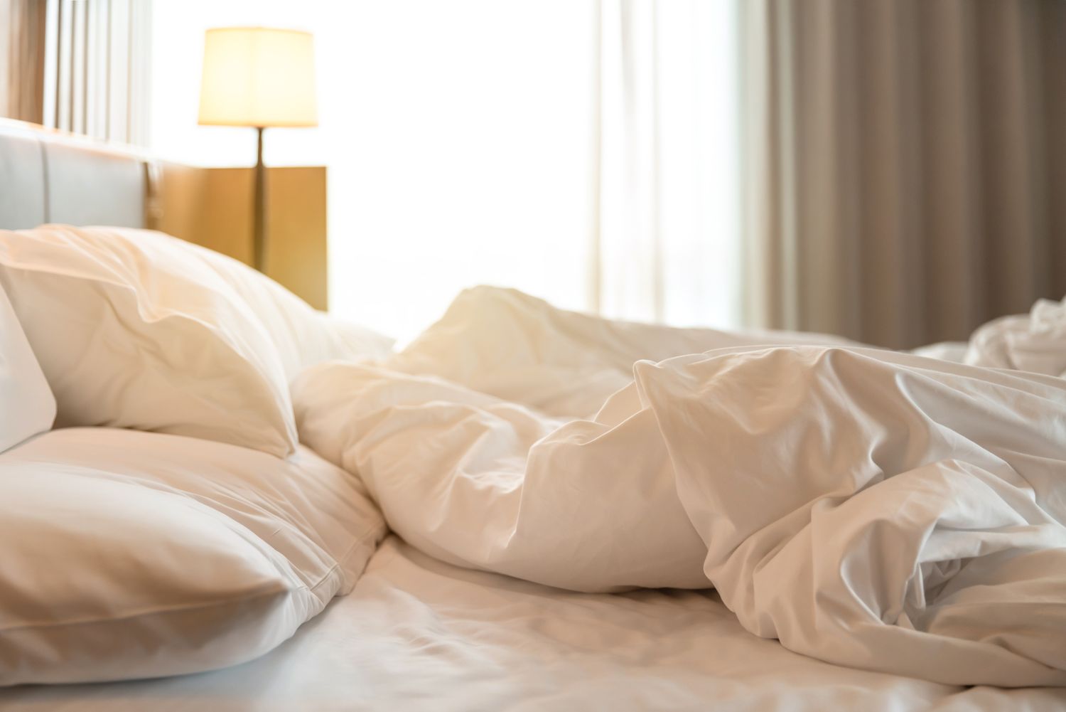 A messy bed with white sheets and pillows a lit bedside lamp in the background