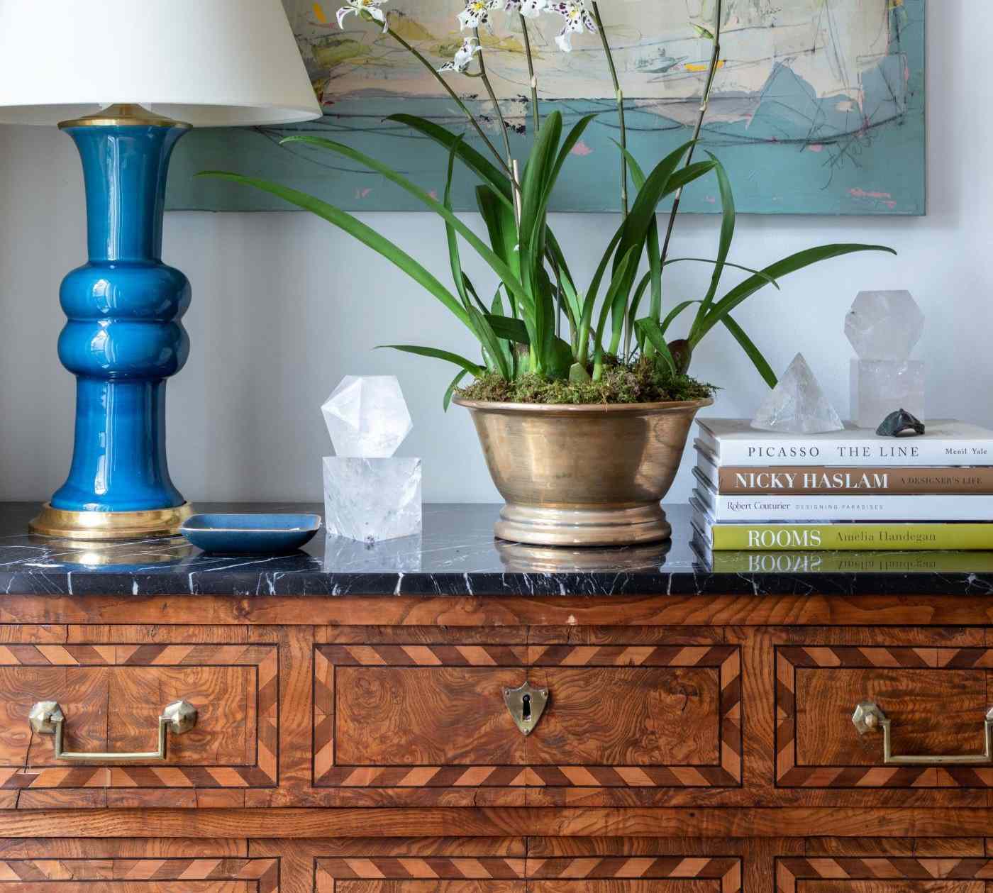 A Louis XVI style chest of drawers topped with a blue lamp potted plant books and decorative items