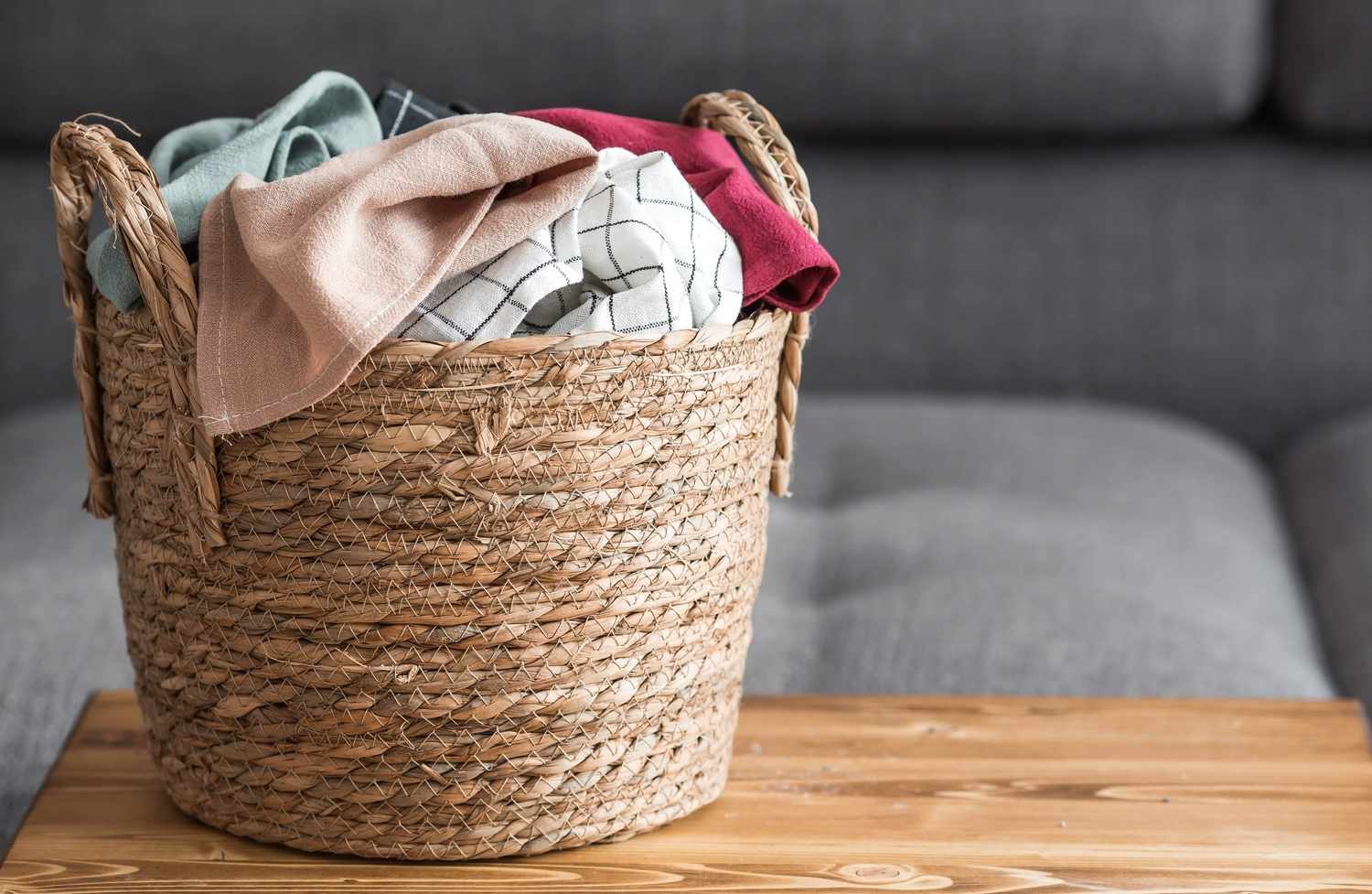 A woven basket containing various clothes placed on a wooden surface with a couch in the background