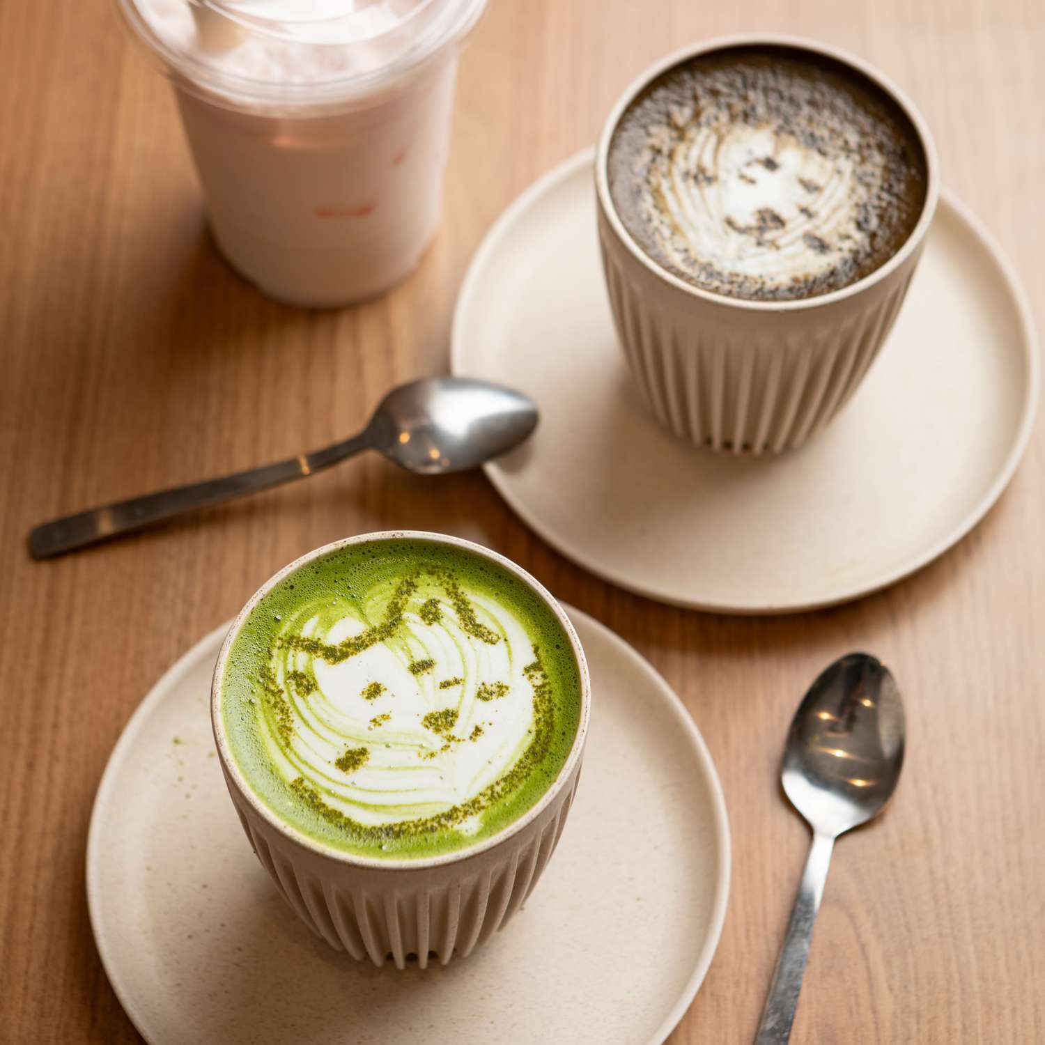 Two ceramic cups of drinks one a matcha beverage and the other a latte placed on saucers on a wooden table with spoons