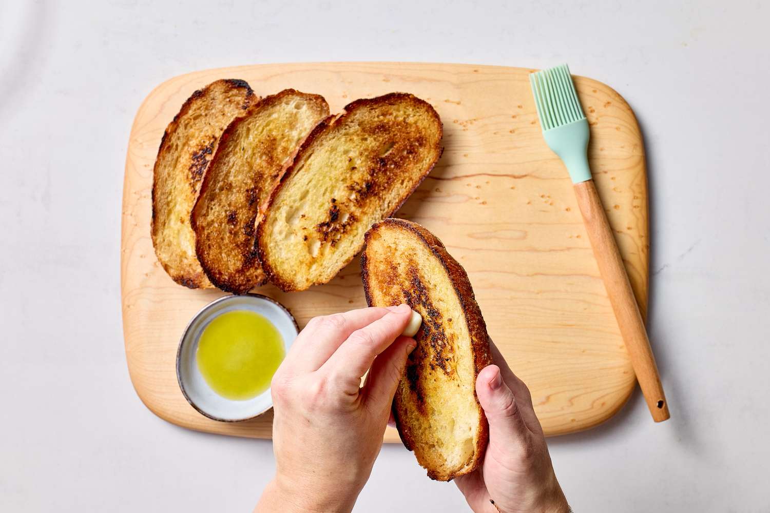 Toasted bread slices being brushed with olive oil on a wooden cutting board