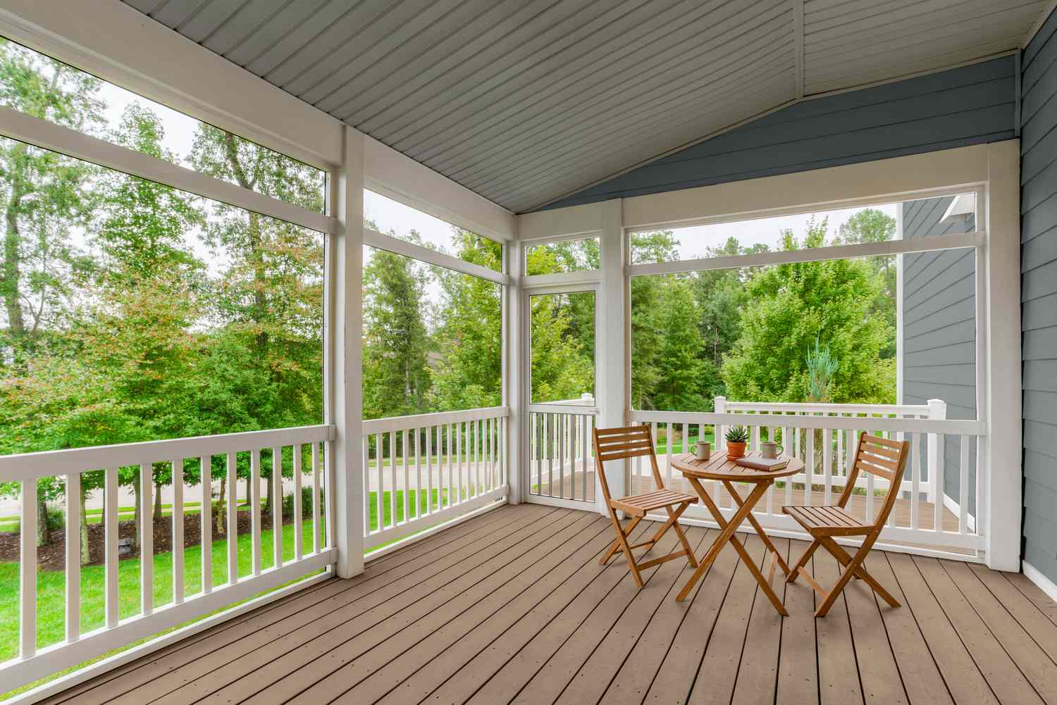 An outdoor wooden deck with two chairs and a small table surrounded by open railings and overlooking a yard with trees