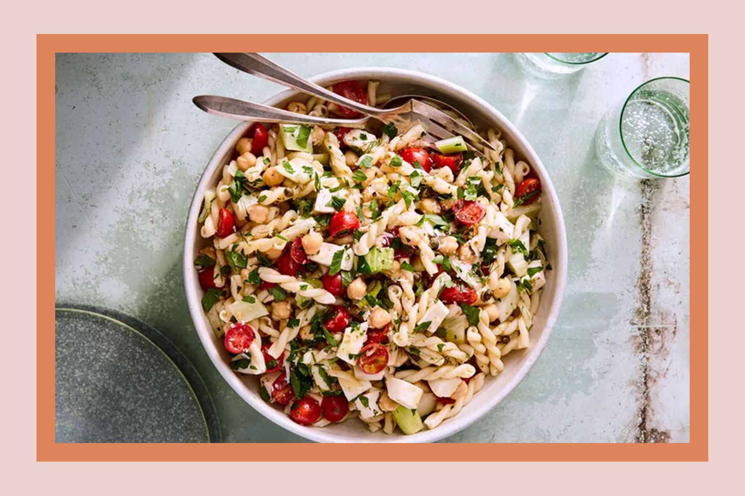 A bowl of pasta salad containing various ingredients such as tomatoes and herbs, with utensils and a glass nearby